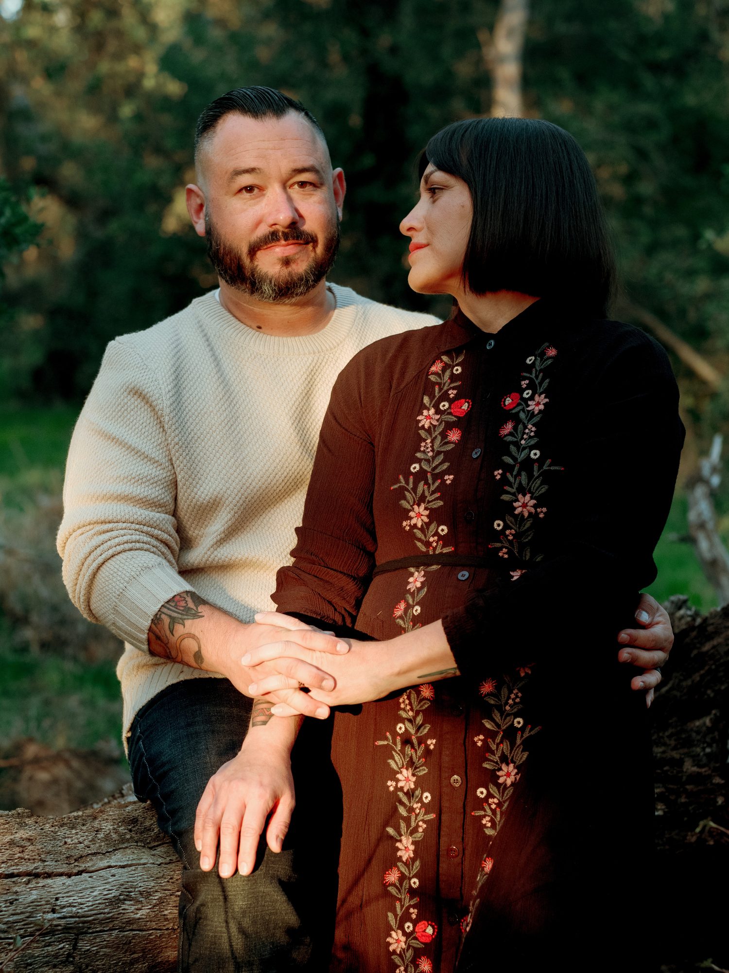 A father and mother sitting together on a log at dusk, she looks at him as he smiles at the camera